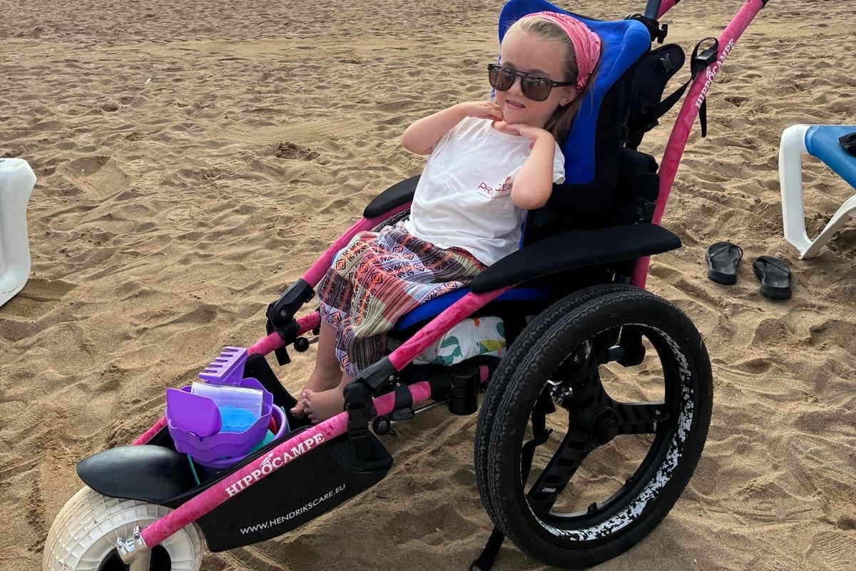 Lanah, kind-ambassadeur van Spieren voor Spieren, zit in een speciale strandrolstoel op het strand en geniet van buiten zijn ondanks haar spierziekte. title=
